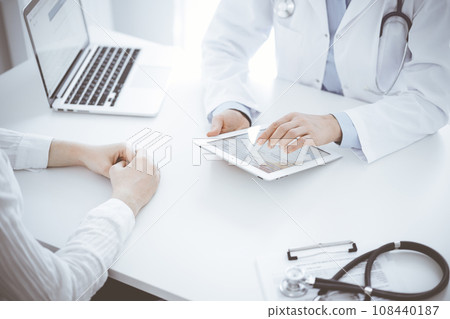 Doctor and patient sitting opposite each other at the desk in clinic. The focus is on female physician's hands pointing into tablet computer touchpad, close up. Medicine concept Doctor and patient sitting opposite each other at the desk in clinic. The focus is on female physician's hands pointing into tablet computer touchpad, close up. Medicine concept 108440187