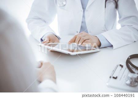 Doctor and patient sitting opposite each other at the desk in clinic. The focus is on female physician's hands using tablet computer, close up. Medicine concept 108440191
