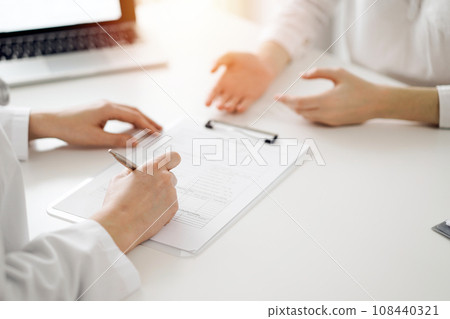 Doctor and patient sitting near each other at the white desk in clinic. Female physician is listening filling up a records form. Medicine concept 108440321