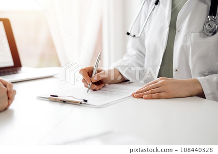 Doctor and patient sitting near each other at the white desk in clinic. Female physician is listening filling up a records form. Medicine concept 108440328