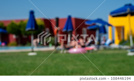 Blurred shot of people relaxing and sunbathing by the pool on sun loungers and under sun umbrellas against the backdrop of bungalows. 108446194
