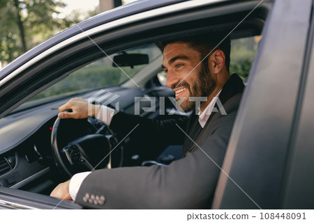 Smiling businessman in suit is riding behind steering wheel of car and looks away 108448091