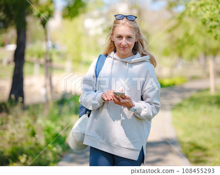 Young beautiful woman using smartphone in city park. Smiling female student texting on mobile phone outdoor. Modern lifestyle, connection, casual business concept 108455293