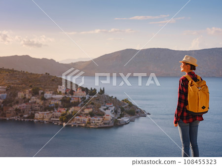 Nice asian Happy Female with backpack Enjoying her holidays on Symi Islands in sunset time. View of port Symi or Simi, is tiny island of Dodecanese, Greece, calm atmosphere and fabulous architecture. Nice asian Happy Female with backpack Enjoying her holidays on Symi Islands in sunset time. View of port Symi or Simi, is tiny island of Dodecanese, Greece, calm atmosphere and fabulous architecture. 108455323