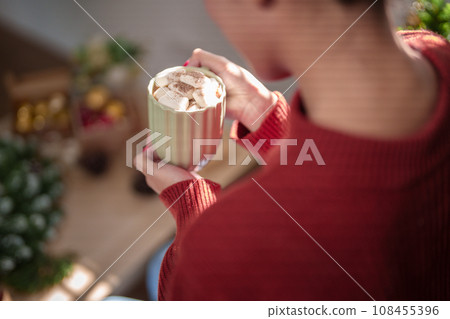 Young asian woman in sweater drinking hot chocolate with marshmallow while sitting near christmas tree in living room decorating ornaments for celebrate christmas festive holiday and winter seasons Young asian woman in sweater drinking hot chocolate with marshmallow while sitting near christmas tree in living room decorating ornaments for celebrate christmas festive holiday and winter seasons 108455396