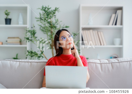 Happy young Asian woman wearing eyeglasses using laptop while seated on couch at home Happy young Asian woman wearing eyeglasses using laptop while seated on couch at home 108455926