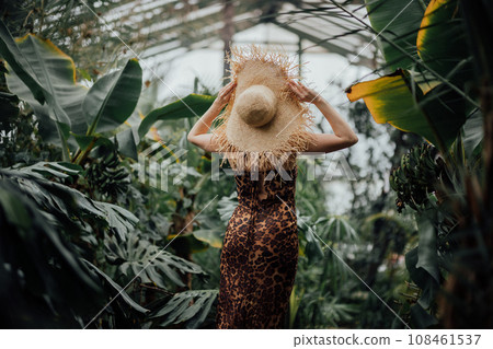 Back view of woman tourist in straw hat and leopard dress standing in palm greenhouse  108461537