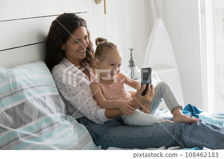Cheerful mother rest on bed with daughter use smartphone Cheerful mother rest on bed with daughter use smartphone 108462183