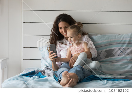 Young mom relaxing in bedroom with daughter use mobile phone Young mom relaxing in bedroom with daughter use mobile phone 108462192