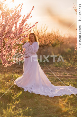 Woman blooming peach orchard. Against the backdrop of a picturesque peach orchard, a woman in a long white dress enjoys a peaceful walk in the park, surrounded by the beauty of nature. Woman blooming peach orchard. Against the backdrop of a picturesque peach orchard, a woman in a long white dress enjoys a peaceful walk in the park, surrounded by the beauty of nature. 108462744