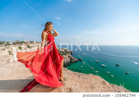 Woman sea red dress yachts. A beautiful woman in a red dress poses on a cliff overlooking the sea on a sunny day. Boats and yachts dot the background. Woman sea red dress yachts. A beautiful woman in a red dress poses on a cliff overlooking the sea on a sunny day. Boats and yachts dot the background. 108462794