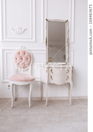 classic room with white patterned wall, dressing table with mirror, decorated with golden and pink soft chair. Details of the interior of the bedroom for girls and make-up, hairstyles with a mirror 108463673
