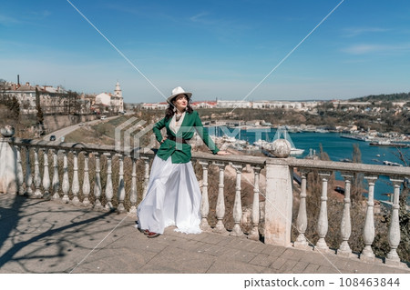 Woman walks around the city, lifestyle. Happy woman in a green jacket, white skirt and hat is sitting on a white fence with balusters overlooking the sea bay and the city. Woman walks around the city, lifestyle. Happy woman in a green jacket, white skirt and hat is sitting on a white fence with balusters overlooking the sea bay and the city. 108463844