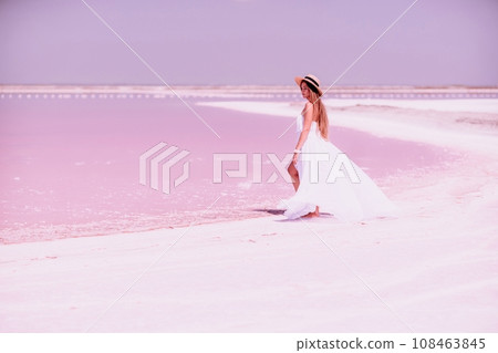 Woman in pink salt lake. She in a white dress and hat enjoys the scenic view of a pink salt lake as she walks along the white, salty shore, creating a lasting memory. Woman in pink salt lake. She in a white dress and hat enjoys the scenic view of a pink salt lake as she walks along the white, salty shore, creating a lasting memory. 108463845
