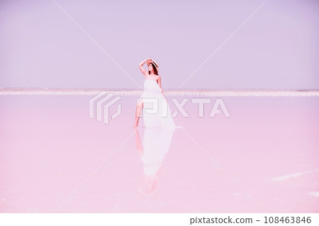 Woman in pink salt lake. She in a white dress and hat enjoys the scenic view of a pink salt lake as she walks along the white, salty shore, creating a lasting memory. Woman in pink salt lake. She in a white dress and hat enjoys the scenic view of a pink salt lake as she walks along the white, salty shore, creating a lasting memory. 108463846
