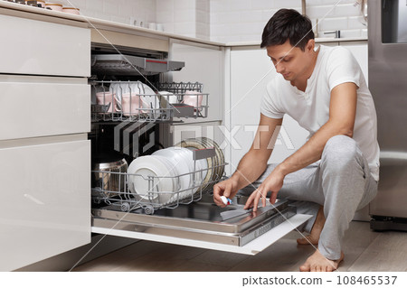 handsome man putting detergent tablet into dishwasher in kitchen handsome man putting detergent tablet into dishwasher in kitchen 108465537