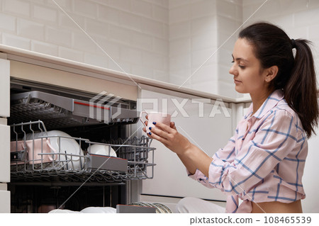 attractive woman unloading cup from open automatic built-in dishwasher machine with clean utensils inside in modern white kitchen. attractive woman unloading cup from open automatic built-in dishwasher machine with clean utensils inside in modern white kitchen. 108465539