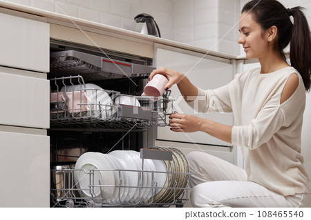 attractive woman unloading cup from open automatic built-in dishwasher machine with clean utensils inside in modern white kitchen. attractive woman unloading cup from open automatic built-in dishwasher machine with clean utensils inside in modern white kitchen. 108465540