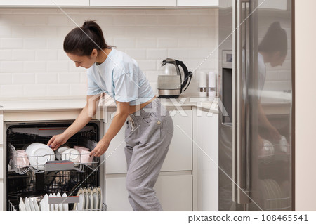 happy woman unloading plate from open automatic built-in dishwasher machine with clean utensils inside in modern white kitchen. 108465541