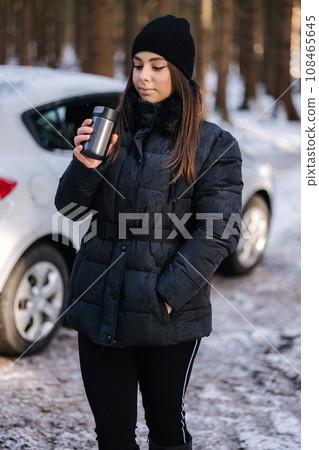 Attractive young woman trink coffee from thermos in front of car. Snowy forest Attractive young woman trink coffee from thermos in front of car. Snowy forest 108465645