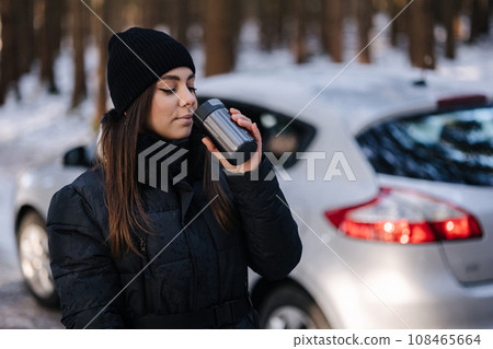 Attractive young woman trink coffee from thermos in front of car. Snowy forest Attractive young woman trink coffee from thermos in front of car. Snowy forest 108465664