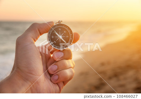 man's hand holds a compass on the beach. Hand with compass over sand by the sea, direction and adventure concept 108466027