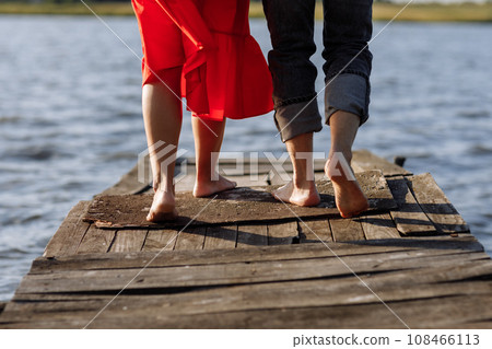 Cropped photo young lovers married couple, husband and wife, held of hands on a wooden bridge near lake. Rear view of couple standing on pier. lower half. Place for text and design. Close Up. 108466113