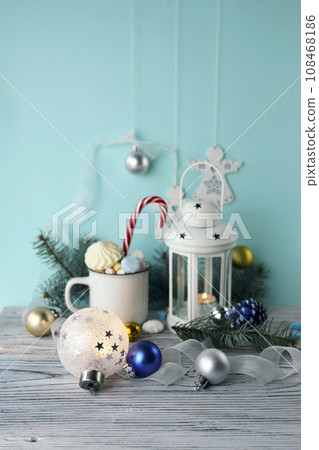 Christmas decorations, a cup of coffee with meringues and marshmallows, a lantern with a burning candle, glowing balls and New Year's toys on a background with fir branches Christmas decorations, a cup of coffee with meringues and marshmallows, a lantern with a burning candle, glowing balls and New Year's toys on a background with fir branches 108468186
