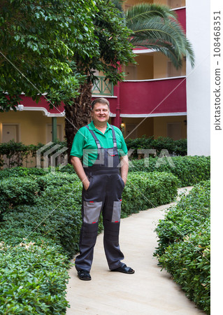 A blond man in work clothes in the green courtyard of a large house 108468351