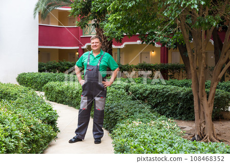 A blond man in work clothes in the green courtyard of a large house A blond man in work clothes in the green courtyard of a large house 108468352