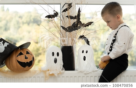 Halloween. Concept. A cute and cheerful boy sits near the window against the background of white ghosts and a vase with dry branches and black paper bats. Halloween. Concept. A cute and cheerful boy sits near the window against the background of white ghosts and a vase with dry branches and black paper bats. 108470856