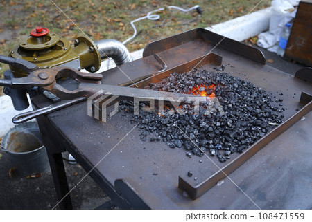 Blacksmith hand holds forceps with a metal billet in a burning forge glowing it red. Smithy workshop Blacksmith hand holds forceps with a metal billet in a burning forge glowing it red. Smithy workshop 108471559