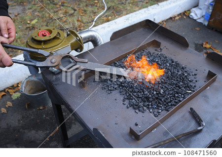 Blacksmith hand holds forceps with a metal billet in a burning forge glowing it red. Smithy workshop 108471560