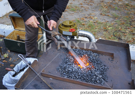 Blacksmith hand holds forceps with a metal billet in a burning forge glowing it red. Smithy workshop 108471561