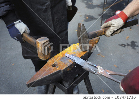 Blacksmith hands holding forceps and a hammer forging a metal billet blade of a knife on an anvil 108471730