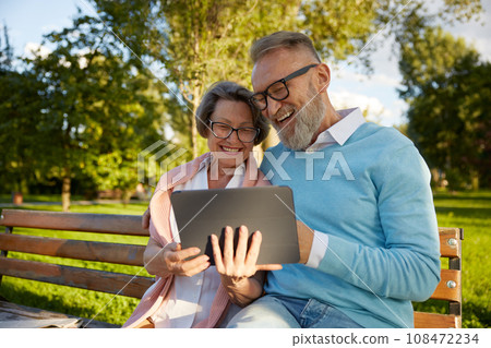 Happy cheerful senior couple using digital tablet while rest on park bench Happy cheerful senior couple using digital tablet while rest on park bench 108472234