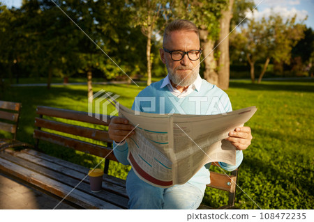 Serious bearded mature man reading daily newspaper on park bench Serious bearded mature man reading daily newspaper on park bench 108472235