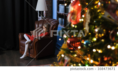 Little girl sitting in comfortable armchair and reading book near Christmas tree at home. Winter holidays and New Year celebration concept Little girl sitting in comfortable armchair and reading book near Christmas tree at home. Winter holidays and New Year celebration concept 108473671