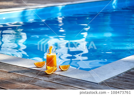Orange juice and sliced oranges near the pool against the background of blue water. Copy space. Front view 108473761