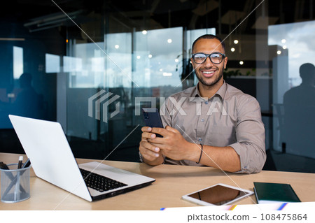 Portrait of a young successful businessman, the man is smiling and looking at the camera, holding a phone in his hands, using an application on a smartphone, working inside the office with a laptop. Portrait of a young successful businessman, the man is smiling and looking at the camera, holding a phone in his hands, using an application on a smartphone, working inside the office with a laptop. 108475864