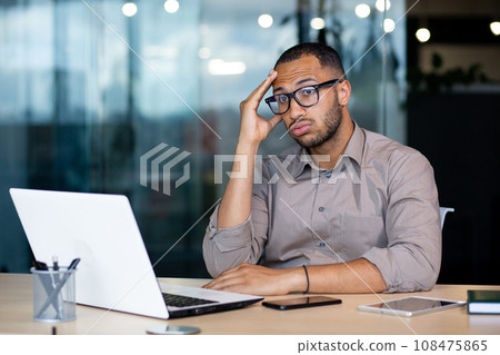 Frustrated and sad man at workplace reading online message from laptop, businessman sitting inside office at workplace, desperate unhappy, got bad news, bankruptcy, layoff. Frustrated and sad man at workplace reading online message from laptop, businessman sitting inside office at workplace, desperate unhappy, got bad news, bankruptcy, layoff. 108475865