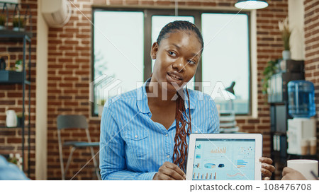 Business analyst explaining statistics, giving presentation in boardroom meeting space with digital tablet. African american team of employees examining online company analytics for development. Business analyst explaining statistics, giving presentation in boardroom meeting space with digital tablet. African american team of employees examining online company analytics for development. 108476708