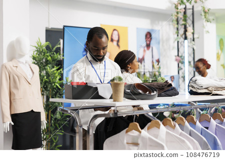 Clothes mall african american worker laying out male shirts and accessories on shelf. Shopping center department man seller working, hanging garment while standing near rack 108476719
