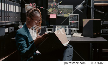 African american policewoman reviewing case files, working on criminal investigation with forensic evidence and suspect photos. Female inspector using detective board in law agency. 108476870