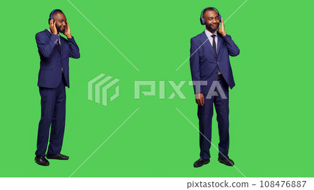 Male manager using headset listening to music on camera, having fun with radio songs over full body green screen. Young businessman in suit dancing and wearing headphones in studio. Male manager using headset listening to music on camera, having fun with radio songs over full body green screen. Young businessman in suit dancing and wearing headphones in studio. 108476887