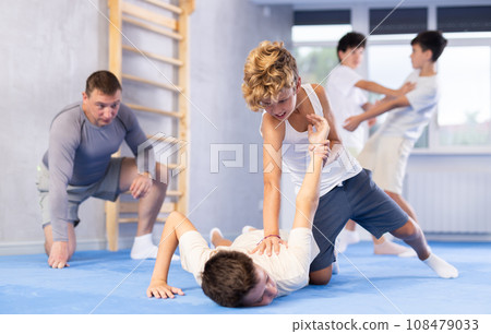 Pair of teenager boys practicing new self-defense moves during training in gym, learning painful pronating wristlock on opponent lying face down on floor Pair of teenager boys practicing new self-defense moves during training in gym, learning painful pronating wristlock on opponent lying face down on floor 108479033