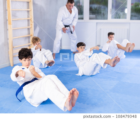Concentrated boys in kimono doing abdominal exercise and practicing punches in gym during group martial arts workout Concentrated boys in kimono doing abdominal exercise and practicing punches in gym during group martial arts workout 108479348