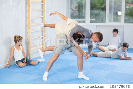 Male karate instructor training little child in dojo or jiu-jitsu in gym. Children sitting on floor around coach 108479678