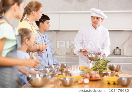 Young guy cook teaches to cook group of children Young guy cook teaches to cook group of children 108479680