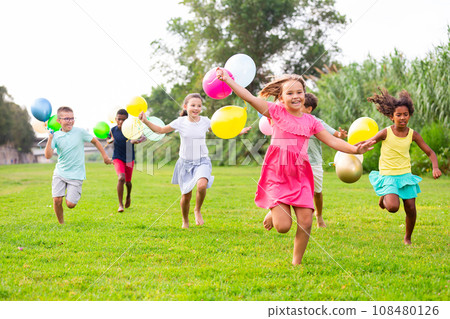 Cheerful preteen boys and girls with balloons running on green lawn 108480126